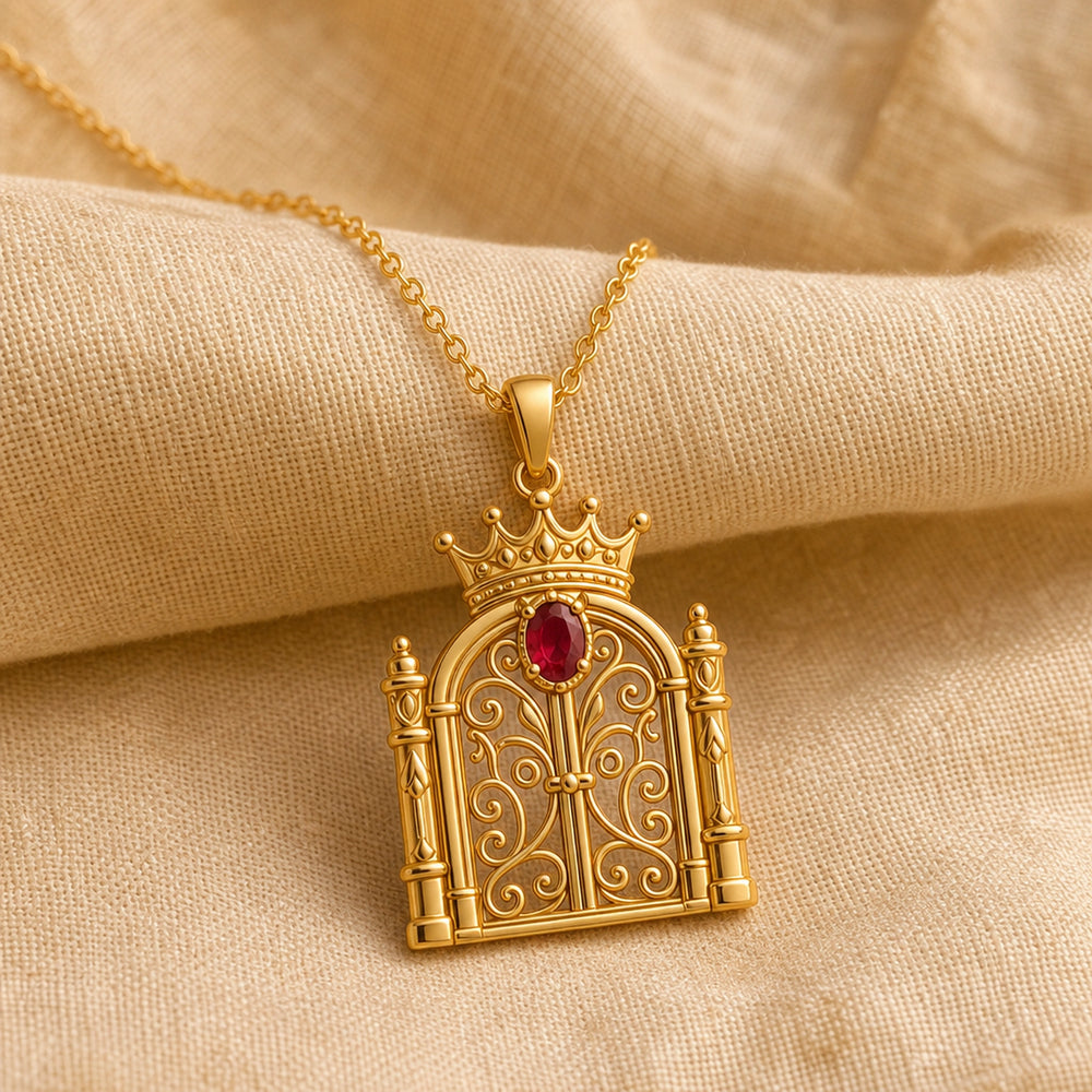 Ruby crowned gate pendant with a deep red oval stone in a raised setting, ornate curling ironwork, framed columns, and a detailed crown above the arched gate. Gold finish.