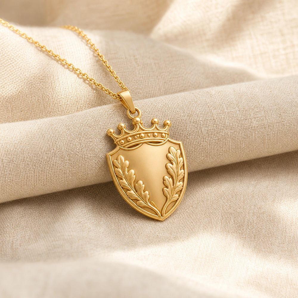 Oakleaf crown shield pendant with a pointed shield shape, raised botanical leaf relief, detailed crown top, smooth center panel, and a polished cable chain. Gold finish.