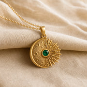 Kepler emerald supernova pendant with a cratered crescent moon, round green emerald center stone in a smooth setting, and detailed radiant starburst rays. Gold finish.