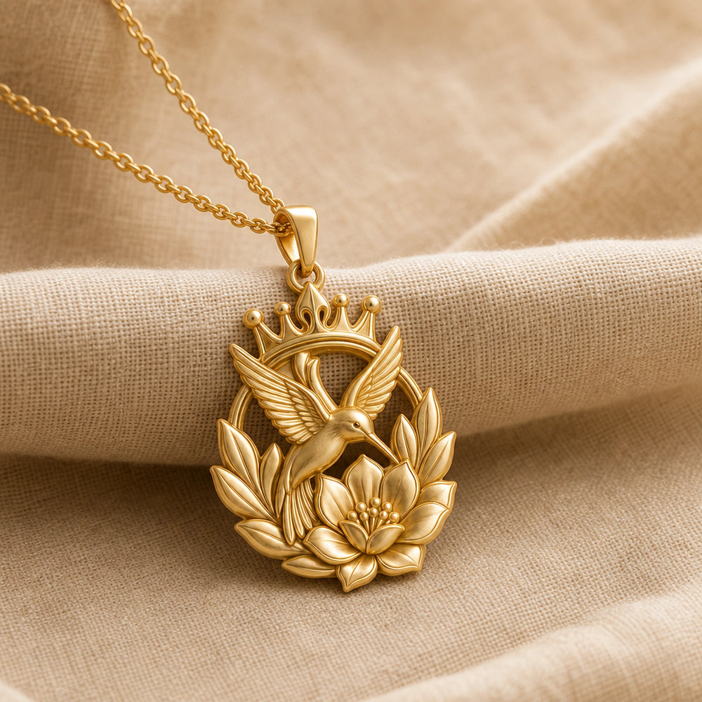 Crownblossom hummingbird pendant necklace with a raised crown, detailed flying bird, open leaf wreath, and sculpted flower in a warm-toned finish. Gold finish.