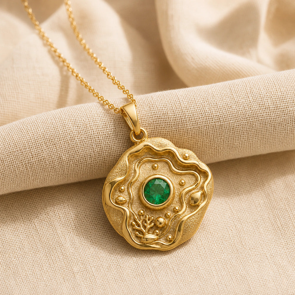 Emerald tide pool pendant necklace with a faceted green center stone in a bezel setting, surrounded by layered wave relief, shell details, coral-inspired accents, and textured ocean motifs. Gold finish.