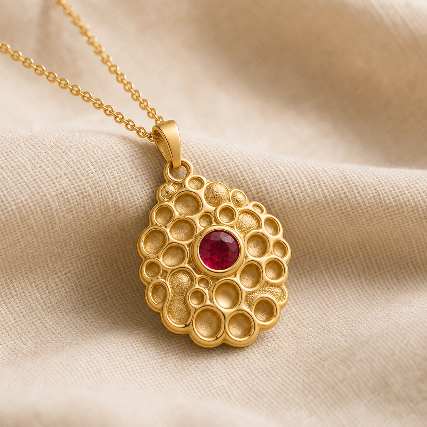 Ruby seafoam pendant with a round deep-red stone in a smooth bezel setting, surrounded by raised textured circular cells in an organic seafoam design on a cable chain. Gold finish.