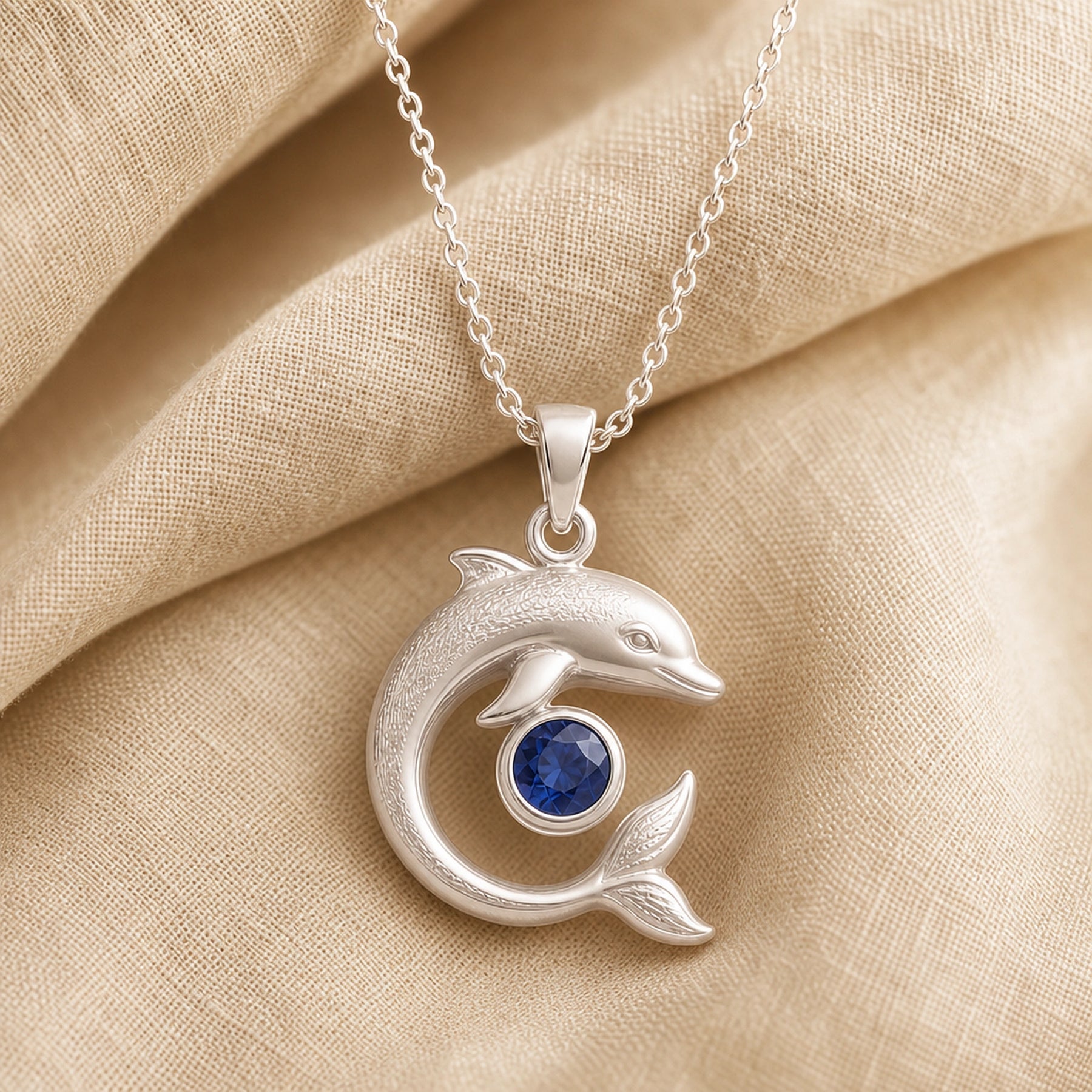 925 sterling silver dolphin pendant in a curved swimming pose around a round blue sapphire centerpiece. Silver finish.