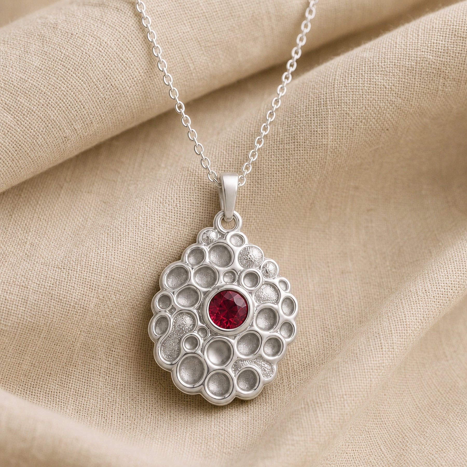 Ruby seafoam pendant with a round deep-red stone in a smooth bezel setting, surrounded by raised textured circular cells in an organic seafoam design on a cable chain. Silver finish.
