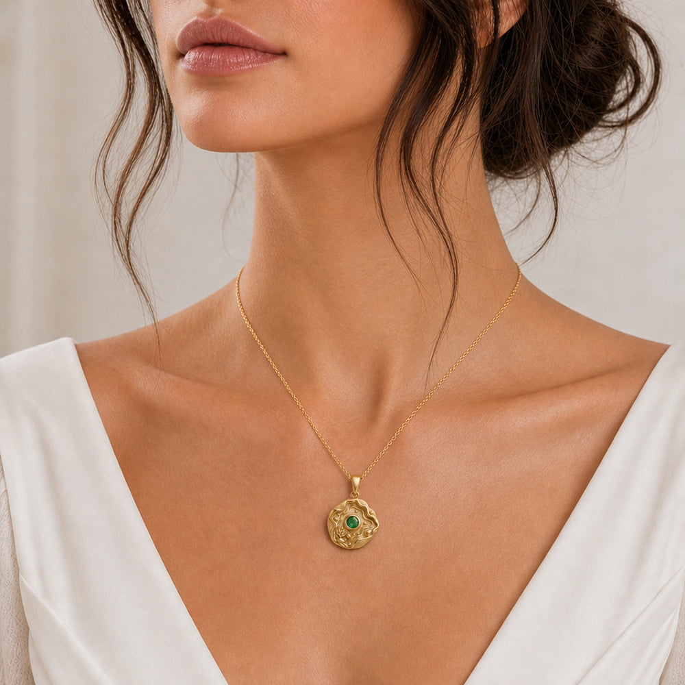 Emerald tide pool pendant necklace with a faceted green center stone in a bezel setting, surrounded by layered wave relief, shell details, coral-inspired accents, and textured ocean motifs. Worn on a model, front view.